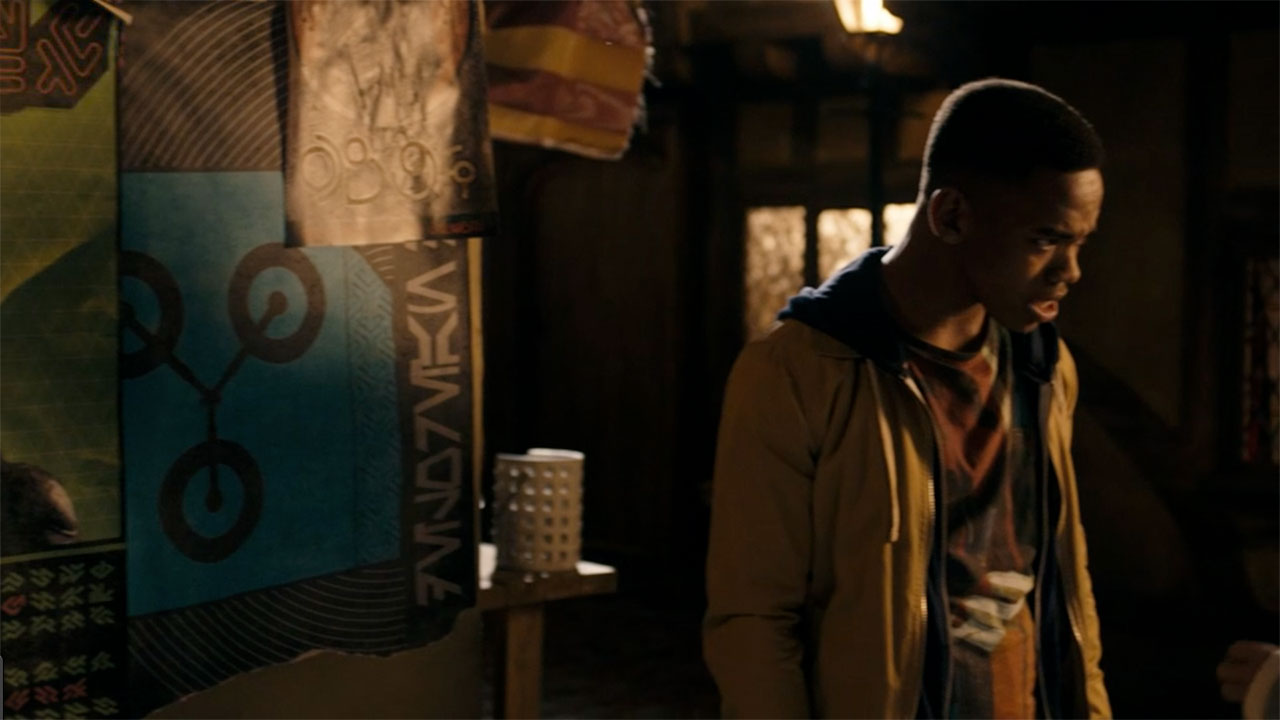 Rigsy (Jovian Wade) stands next to a wall with posters on it, one of which depicts a flux capacitor with "DeLorean" written in Aurebesh